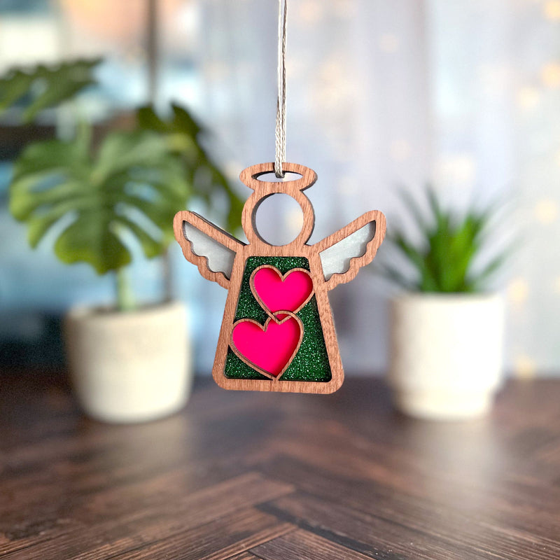The Forged Flare® Limited Edition Interlocking Hearts Ornament – Series 8 features a 3.5" wooden angel figurine with green glitter, hanging by a string. Blurred potted plants and soft lights create a cozy backdrop on a wooden surface.