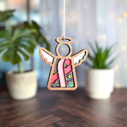 The Forged Flare® Mother's Angels® 3.5" Breast Cancer Awareness Ribbon for Charity Ornament features a wooden angel with a pink ribbon and colorful beads, making it a thoughtful gift amid softly-lit greenery in the background.