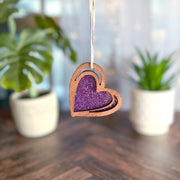 The Forged Flare® "I Love You" Purple Heart Ornament, 2.5" (H), features a glittery center and sketch-style design, hanging by a string with two blurred potted plants in the background on a wooden surface.