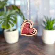 The 2.5" "I Love You" Red Heart Ornament by Forged Flare® hangs on a string, with two potted plants softly blurred in the background on a wooden surface.