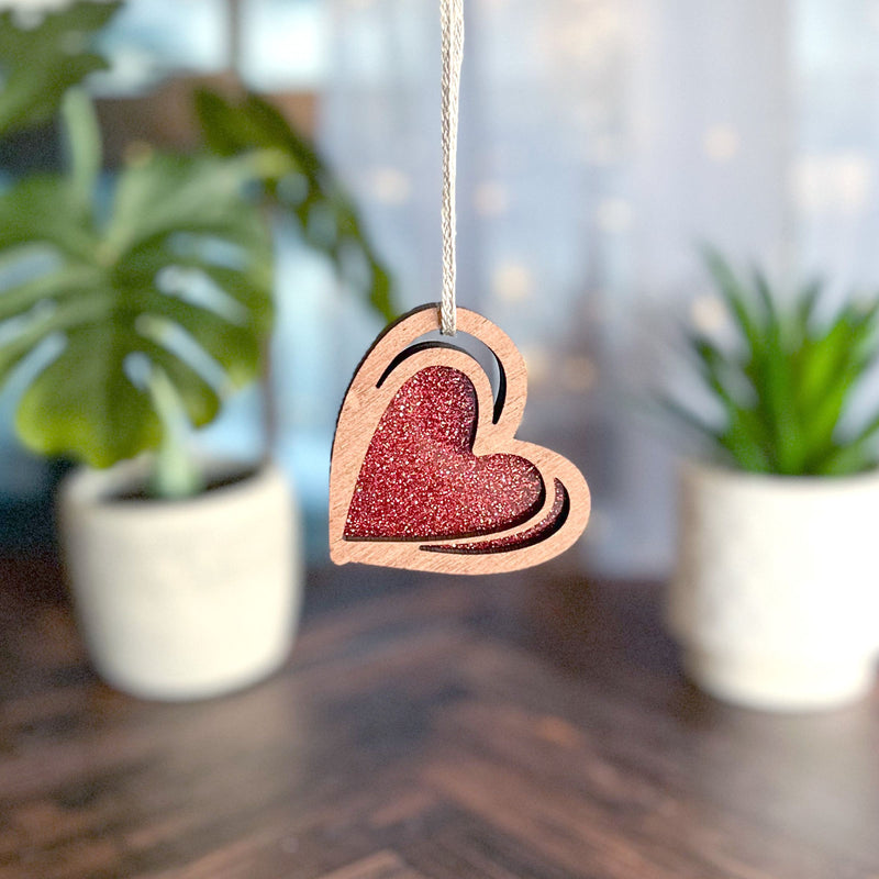The 2.5" "I Love You" Red Heart Ornament by Forged Flare® hangs on a string, with two potted plants softly blurred in the background on a wooden surface.