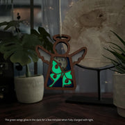 The Forged Flare® Luna Moth Ornament (3.5" Angel Figurine, Mother's Angels®) hangs by a string, its glow-in-the-dark green wings shining after exposure to light. Potted plants and a large translucent stone appear in the background.