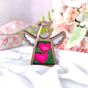 The Forged Flare® Limited Edition Interlocking Hearts Ornament - Series 8 is a 3.5" Mother's Angels® figurine with white wings and interlocking hearts, displayed on a white cloth with pink flowers and heart-themed ribbon in the background.