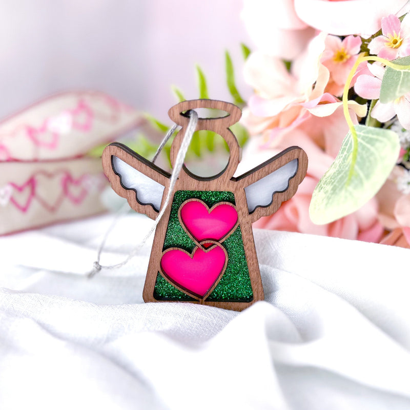 The Forged Flare® Limited Edition Interlocking Hearts Ornament - Series 8 is a 3.5" Mother's Angels® figurine with white wings and interlocking hearts, displayed on a white cloth with pink flowers and heart-themed ribbon in the background.