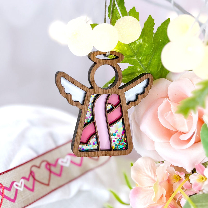 The Forged Flare® Mother's Angels® 3.5" Breast Cancer Awareness Ribbon for Charity Ornament features a wooden angel with a pink ribbon and glitter, perfect as a thoughtful breast cancer gift displayed amid green leaves and pink flowers.