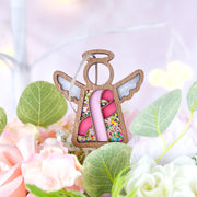 The Breast Cancer Awareness Ribbon for Charity Ornament by Forged Flare® is a 3.5" wooden angel figurine with a halo, wings, and pink ribbon—an ideal gift from Mother's Angels®, set among green leaves and soft pink flowers on a pastel background.