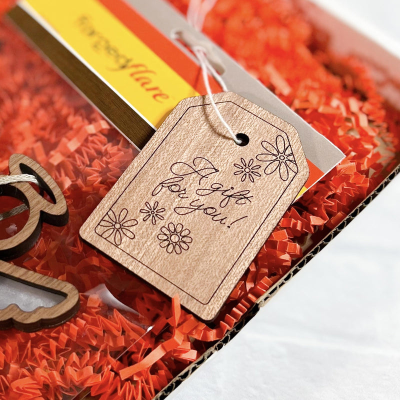 A Forged Flare® Gift Tag—"A gift for you!", 3" (H)—with floral designs adds a charming message, displayed on shredded orange paper inside a gift box.