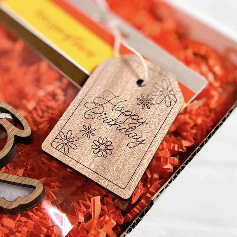 A Forged Flare® "Happy Birthday" gift tag (3" H) with flower designs decorates a package filled with orange crinkle paper.