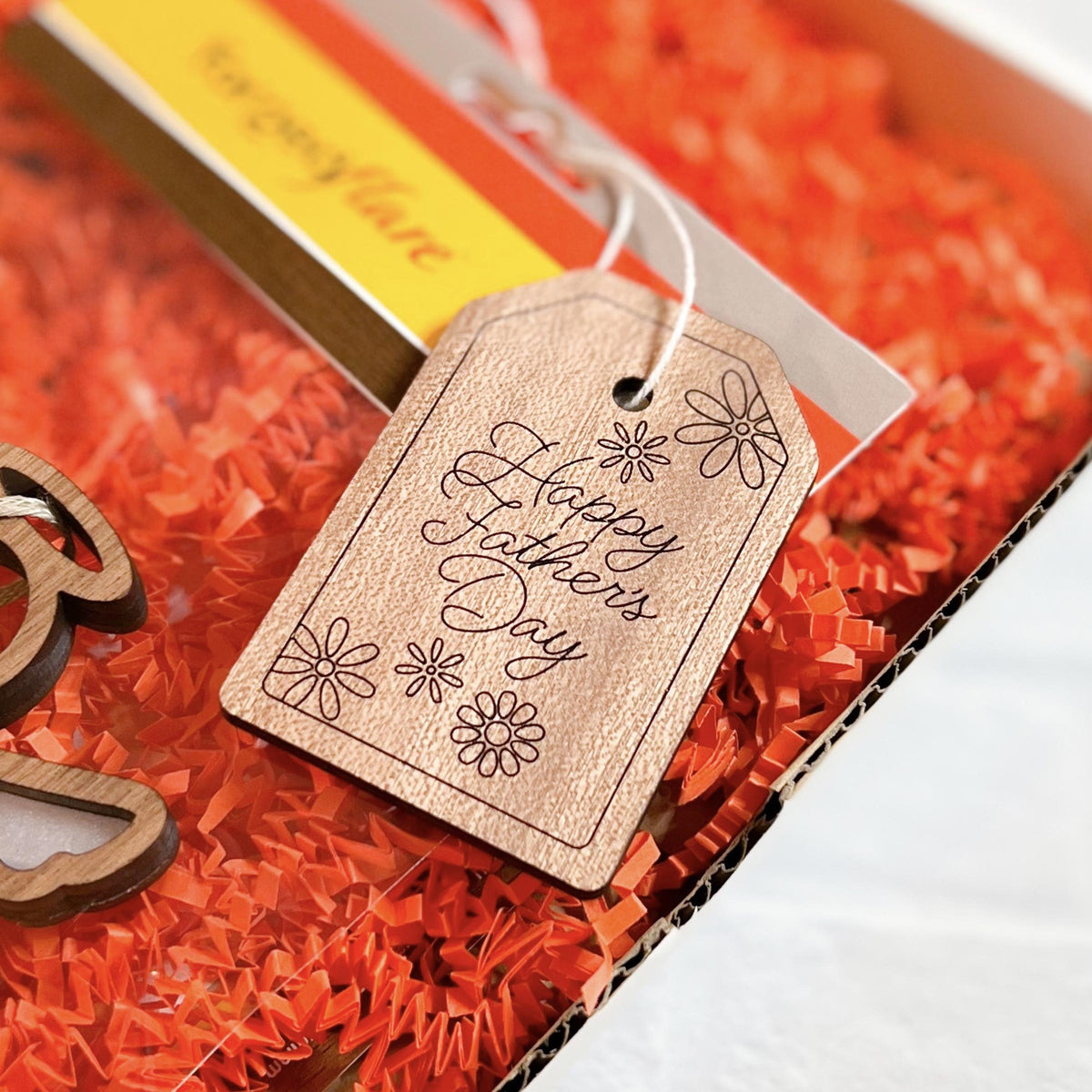 A close-up of the Forged Flare® 3" "Happy Father's Day" Gift Tag with floral design, attached to a present filled with orange shredded paper.