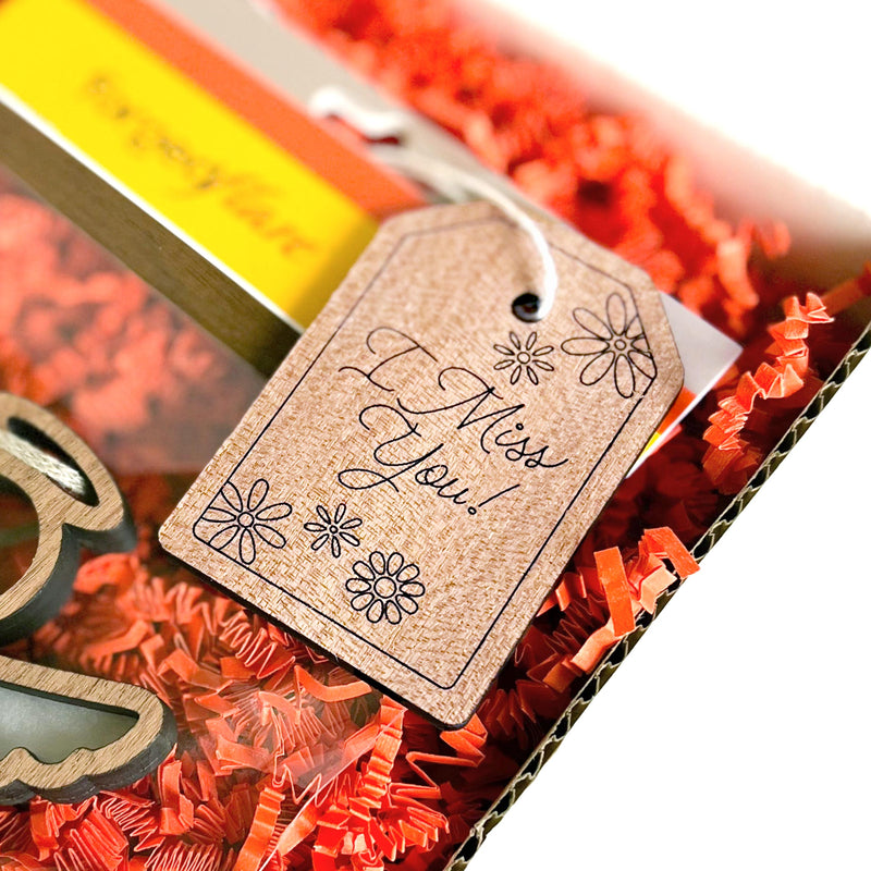 A cardboard gift box with orange paper shreds features a Forged Flare® Gift Tag - "I Miss You!", 3" (H), decorated with flowers and a handwritten message.