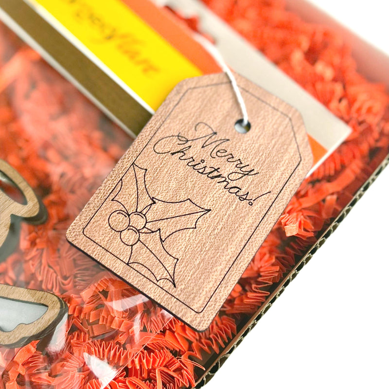 The Forged Flare® Gift Tag - "Merry Christmas!", 3" (H), featuring a holly illustration, is displayed on red shredded paper inside a box.