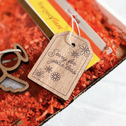 The Forged Flare® 3" wooden gift tag with “Sorry for your loss.” rests on orange shredded paper next to a small wooden angel and a yellow card in a condolence gift box.