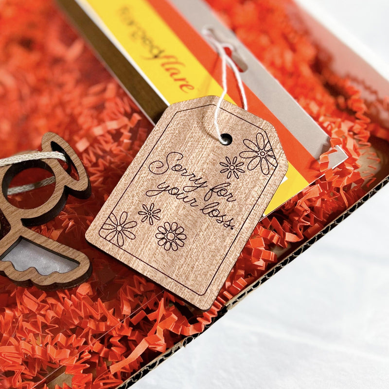 The Forged Flare® 3" wooden gift tag with “Sorry for your loss.” rests on orange shredded paper next to a small wooden angel and a yellow card in a condolence gift box.