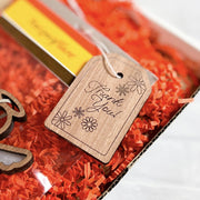 A "Thank You!" gift tag from Forged Flare®, featuring intricate floral designs etched onto sturdy wood and tied with white string, adorns a gift presented with red crinkle paper and an angel ornament. This 3-inch tag beautifully conveys a sense of gratitude.