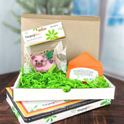 A cardboard box filled with green shredded paper contains a vibrant flying pig-shaped keychain sealed in a clear package, along with an orange Custom Gift Message card by Wrapped. A tree is softly blurred in the background.