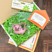 A gift box featuring green paper shreds, a flying pig-themed wooden magnet, and a small orange envelope for your Custom Gift Message. The packaging showcases the brand name "Wrapped," set against a wooden surface backdrop.