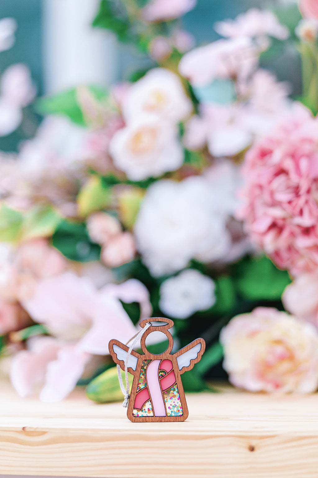 A wooden angel ornament with a pink ribbon stands before soft floral blooms, a gentle nod to breast cancer awareness.