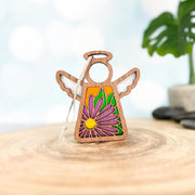 The Forged Flare® Mother's Angels® Aster Ornament is a 3.5" wooden angel figurine with white wings and a purple aster flower design, shown on wood with an orange backdrop and a blurred plant in the distance.