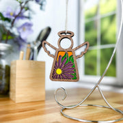 The Forged Flare® Mother's Angels® Aster Ornament, a 3.5" wooden angel with stained-glass-style wings and a purple aster September birth flower, hangs on a metal stand on a wooden desk by a bright window.