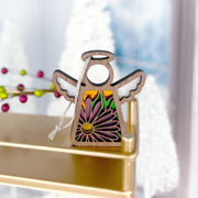 The Forged Flare® Aster Ornament | 3.5" (H) Angel Figurine from Mother's Angels® features cut-out wings and purple aster designs for September, displayed on a gold shelf with snow-covered trees and red berries softly blurred behind.