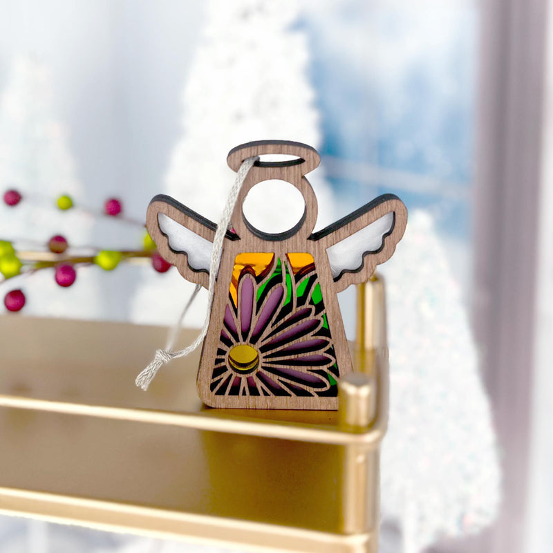 The Forged Flare® Aster Ornament | 3.5" (H) Angel Figurine from Mother's Angels® features cut-out wings and purple aster designs for September, displayed on a gold shelf with snow-covered trees and red berries softly blurred behind.