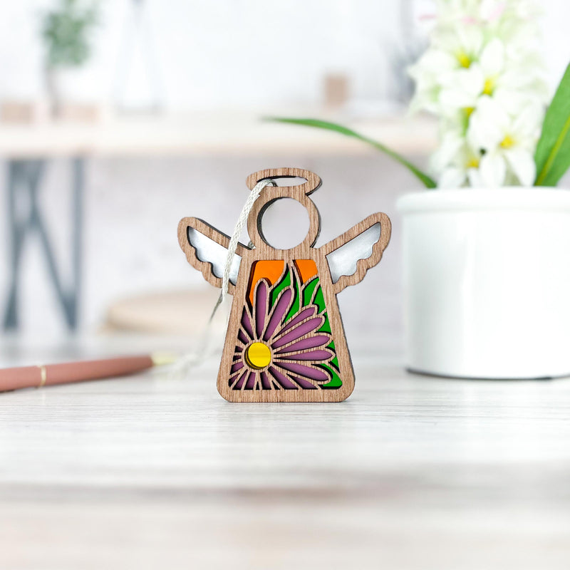 The Forged Flare® Aster Ornament | 3.5" (H) Angel Figurine from Mother's Angels® stands on a light wooden table, showcasing a cutout purple aster flower for September. In the background are white flowers in a pot and blurred office items.