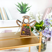 The Forged Flare® Mother's Angels® Aster Ornament, a 3.5" angel figurine, stands on a gold shelf, while a purple September birth flower adds charm among jars and books on cozy white shelves.