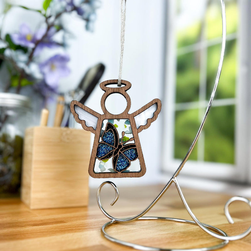 A Forged Flare® Mother’s Angels® 3.5" angel ornament with a Morpho blue butterfly hangs from a silver stand on a wooden table, softly backed by blurred flowers and a window. Warehouse Return - Blue Butterfly Angel Figurine.