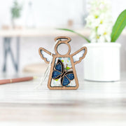A Forged Flare® Mother's Angels® 3.5" Morpho Blue Butterfly Ornament, a wooden angel figurine, sits on a light surface with a white hanging string. The softly blurred background features a white potted plant and decor.