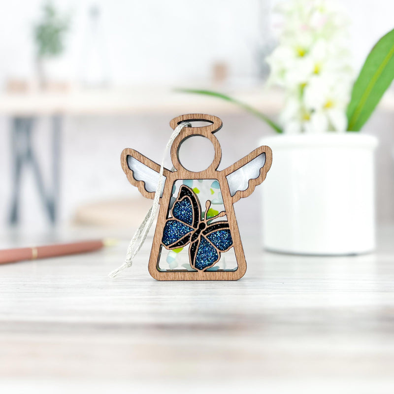 A Forged Flare® Mother's Angels® 3.5" Morpho Blue Butterfly Ornament, a wooden angel figurine, sits on a light surface with a white hanging string. The softly blurred background features a white potted plant and decor.