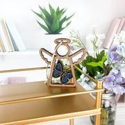The Forged Flare® Mother's Angels® Morpho Blue Butterfly Ornament (3.5") from the Warehouse Return collection stands on a gold shelf, with books, white shelves, and vases of white and purple flowers in the background.