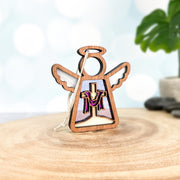 Introducing the Limited Edition By Grace Ornament, a 3.5" angel figurine by Mother's Angels® from Forged Flare®. This beautifully crafted wooden ornament features an angel with outstretched wings and a halo. Inside the angel's body, you'll find a delicately carved cross adorned with a draped purple cloth. Perfect as an Easter gift or decoration, the ornament rests on a round wooden base and is set against a soft-focus backdrop featuring a potted plant and stones.