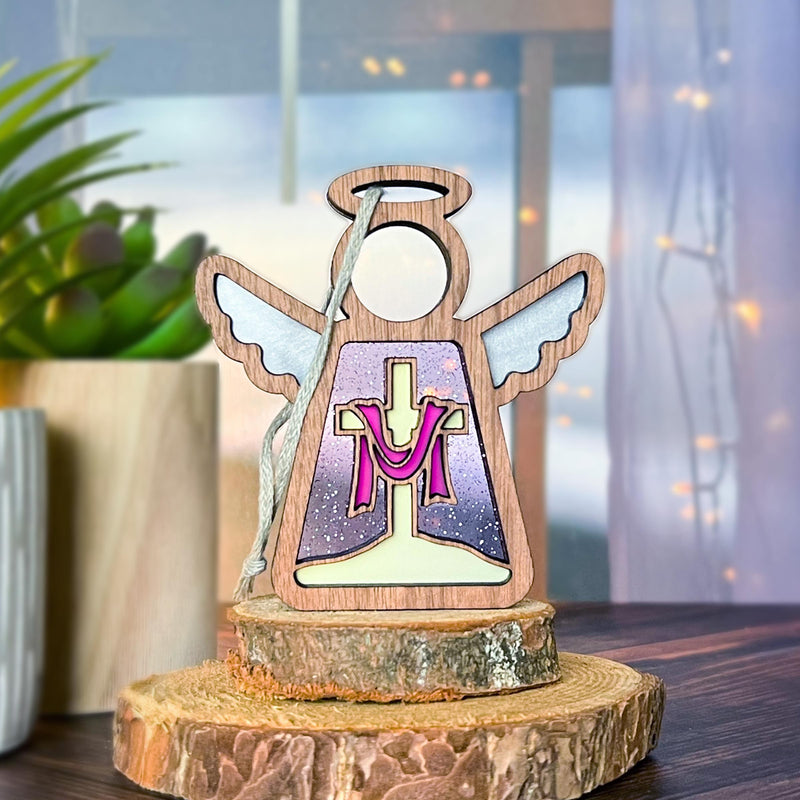The Limited Edition By Grace Ornament from Forged Flare® features a 3.5" angel figurine with a halo and wings, elegantly posed on a stump. The front of the Mother's Angels® figurine is adorned with a cross draped in purple cloth, making it an ideal Easter gift. In the background, there is a blurred view of a plant and window accented with string lights.