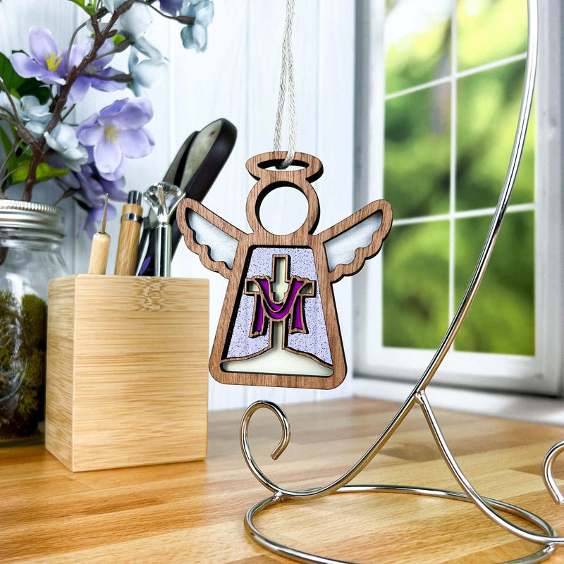The Limited Edition By Grace Ornament, a 3.5" angel figurine from Forged Flare's Mother's Angels® collection, hangs gracefully on a metal stand. Its wooden body features an inset cross with purple and white accents, making it an ideal Easter decoration. In the background, a window offers a view of greenery alongside a vase of purple flowers resting on a wooden counter.