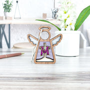 The Limited Edition By Grace Ornament by Forged Flare® is a 3.5" wooden Mother's Angels® figurine with a purple and gold "M," perfect for Easter decor. It stands on a light desk near a white potted plant and pen.