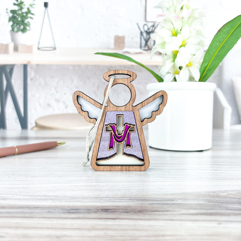 The Limited Edition By Grace Ornament by Forged Flare® is a 3.5" wooden Mother's Angels® figurine with a purple and gold "M," perfect for Easter decor. It stands on a light desk near a white potted plant and pen.