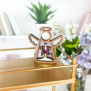 A Limited Edition By Grace Ornament 3.5" Angel Figurine from the Mother's Angels® collection by Forged Flare® stands on a gold metallic shelf. Behind it are books and a vase of flowers, including white and purple blooms. The angel figurine features a small cutout and a violet design on its robe, creating a charming and serene setting—perfect as an Easter gift.