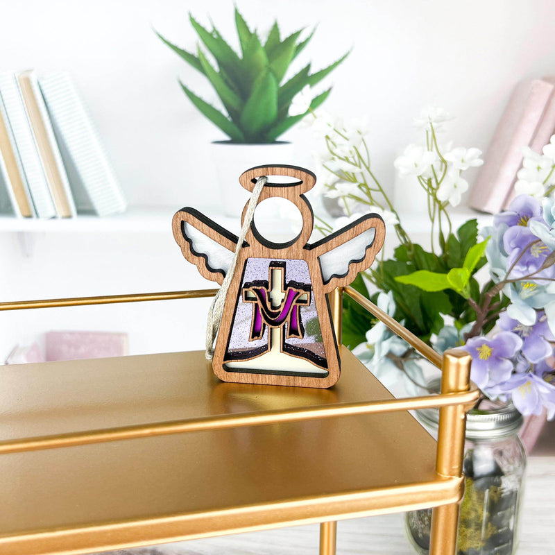 A Limited Edition By Grace Ornament 3.5" Angel Figurine from the Mother's Angels® collection by Forged Flare® stands on a gold metallic shelf. Behind it are books and a vase of flowers, including white and purple blooms. The angel figurine features a small cutout and a violet design on its robe, creating a charming and serene setting—perfect as an Easter gift.