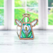 The Forged Flare® Mother's Angels® Calla Lily Ornament is a 3.5" stained glass angel figurine with a wooden outline and hanging string, shown against a white surface with blurred green and white windows in the background.