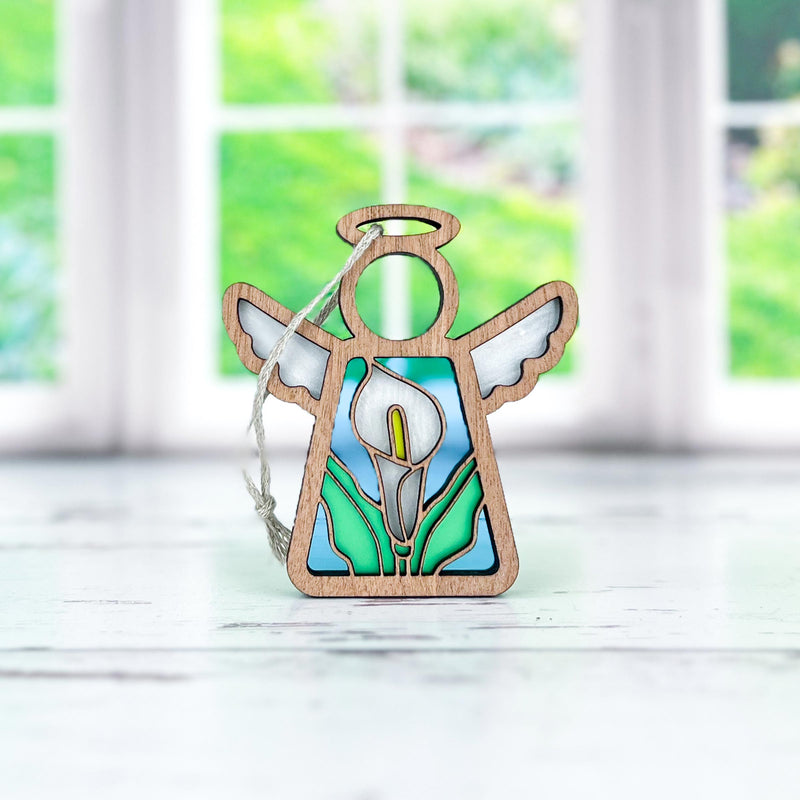 The Forged Flare® Mother's Angels® Calla Lily Ornament is a 3.5" stained glass angel figurine with a wooden outline and hanging string, shown against a white surface with blurred green and white windows in the background.