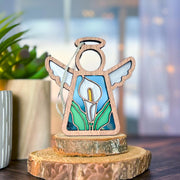 The Forged Flare® Calla Lily Ornament | 3.5" (H) Angel Figurine from the Mother's Angels® line features a stained-glass-style design, standing on a wooden slab amid potted plants—a unique Christmas tree ornament for any setting.