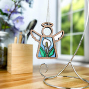 The Forged Flare® Calla Lily Ornament, a 3.5" Mother's Angels® angel figurine, hangs from a silver stand on a wooden desk with blurred flowers, a pen holder, and sunlight streaming through the window in the background.