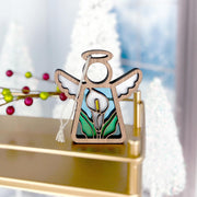 The Forged Flare® Mother's Angels® Calla Lily Ornament is a 3.5" wooden angel figurine with white wings, a halo, and stained glass calla lily details, displayed on a gold shelf surrounded by blurred holiday decor.