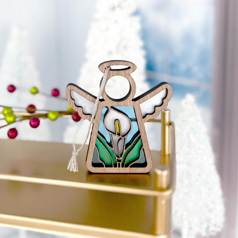 The Forged Flare® Mother's Angels® Calla Lily Ornament is a 3.5" wooden angel figurine with white wings, a halo, and stained glass calla lily details, displayed on a gold shelf surrounded by blurred holiday decor.
