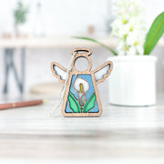 The Forged Flare® Mother's Angels® Calla Lily Ornament is a 3.5" tall wooden angel figurine with a stained glass-style calla lily design, pictured on a light wooden surface with a blurred plant and desk in the background.
