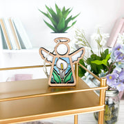The Forged Flare® Calla Lily Ornament | 3.5" (H) Mother's Angels® figurine features a stained glass-style angel with floral design—ideal as a unique Christmas tree ornament or gift. Displayed on a gold shelf with books, vases, and a green plant nearby.