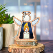 The Forged Flare® Burning Candle Ornament—a 3.5" Mother's Angels® figurine—features a stained glass candle design, perfect as a Christmas angel ornament or collectible, displayed on wood beside a potted plant with a softly blurred background.