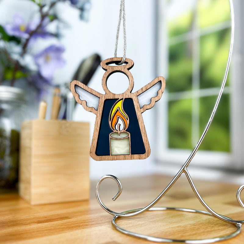 The Forged Flare® Mother's Angels® Burning Candle Ornament is a 3.5" wooden angel figurine with halo and wings, displayed on a metal stand near a window, flowers, and a pen holder on a desk.