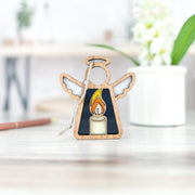 The Forged Flare® Burning Candle Ornament is a 3.5" wooden Mother's Angels® figurine with a stained glass candle design, perfect as a Christmas or memorial gift, shown on light wood with a white pot and pencil blurred in the background.