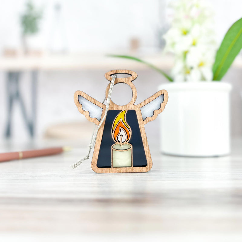 The Forged Flare® Burning Candle Ornament is a 3.5" wooden Mother's Angels® figurine with a stained glass candle design, perfect as a Christmas or memorial gift, shown on light wood with a white pot and pencil blurred in the background.